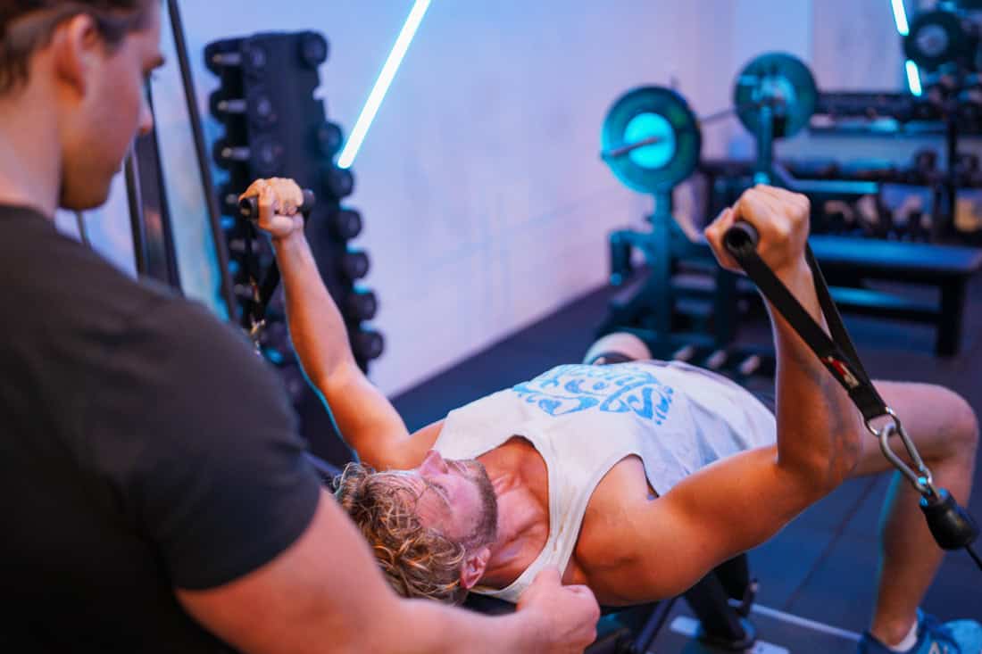 Man ligt op een trainingsbank met kabelgrepen in beide handen terwijl een trainer meekijkt