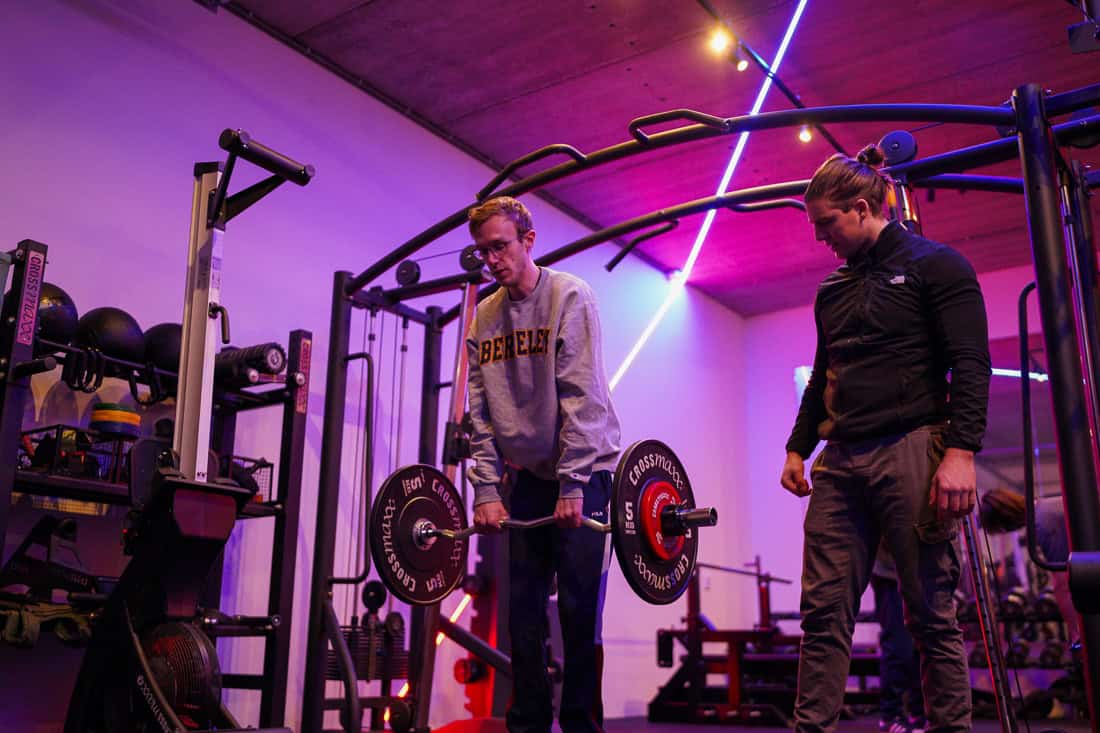 Man houdt een EZ-bar vast met zijn bovenlichaam naar voren gekanteld terwijl een trainer meekijkt