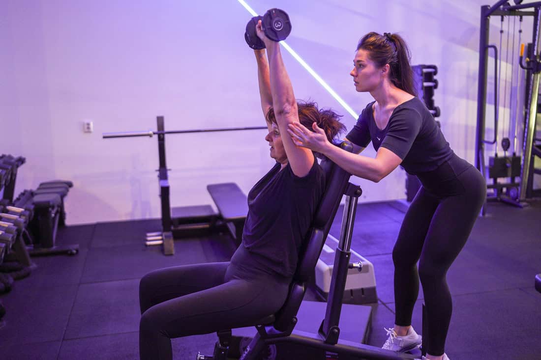 Vrouw doet een seated dumbbell shoulder press met begeleiding