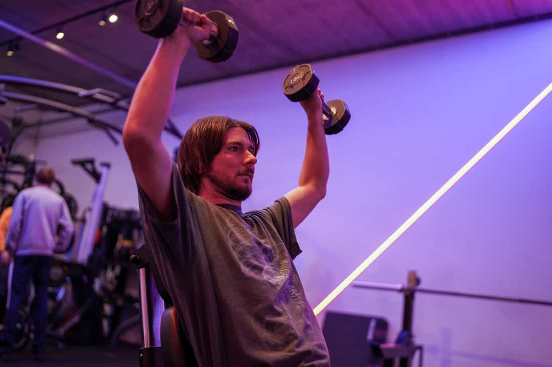 Zittende shoulder press waarbij de dumbbells boven het hoofd worden uitgeduwd