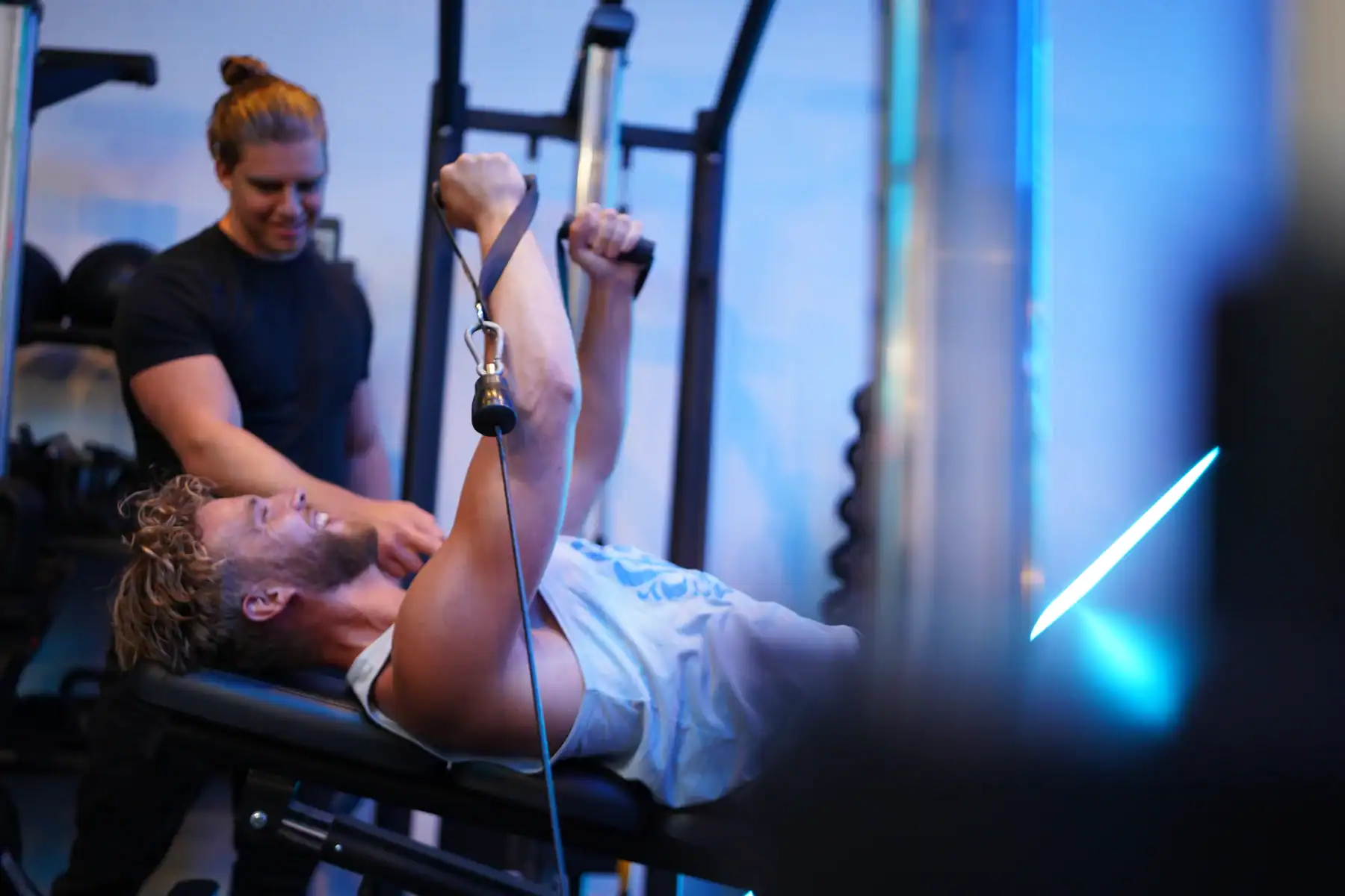 Een personal trainer begeleidt een man bij een oefening met kabels op een trainingsbank.